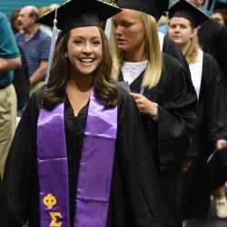 A line of students march in a Commencement ceremony, where the student in the front of the line smiles toward the camera in a cap and gown